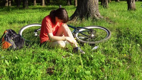 A young woman experiences pain in the knee joint, she blows on the bleeding leg wound after riding a bicycle. The girl is very hurt, the tourist is swinging sitting in the grass in the forest.