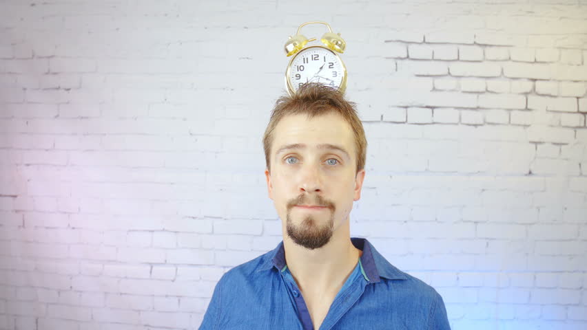 Person showing a ticking clock on head in slow motion 4K. Wide lens portrait of a man in focus pointing finger towards the clock on the head. Bricks wall in the background.