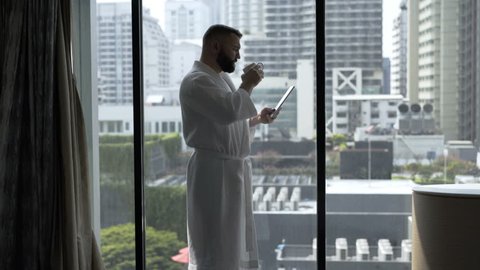 Man in bathrobe drinks coffee and uses tablet in his hotel room 
