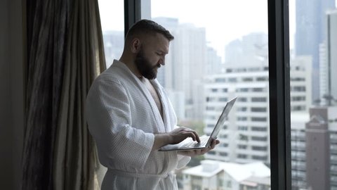 Man wearing bathrobe and working on laptop next to the window in his hotel room 
