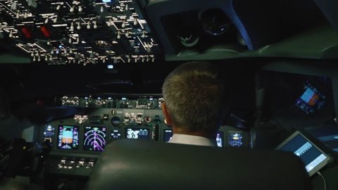Captain is controls the airplane at night, rear view. Pilot in cockpit of passenger airliner.