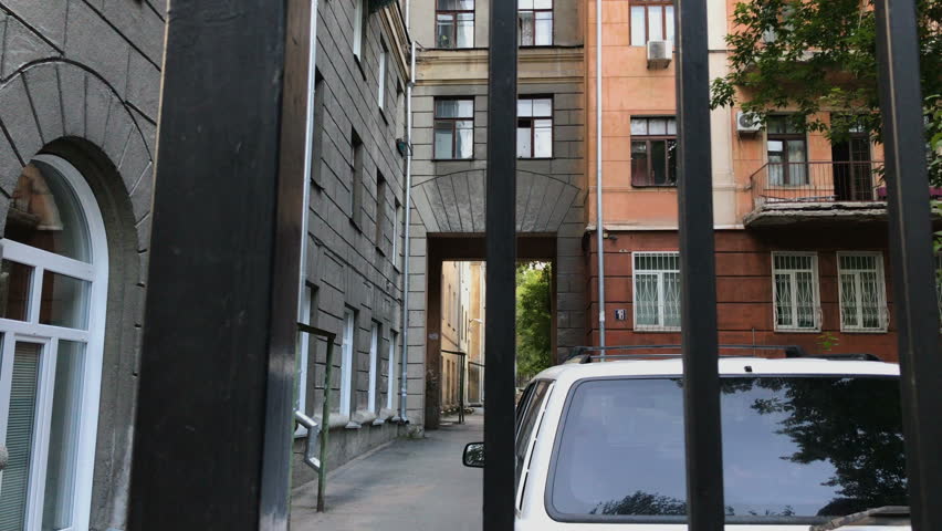 A moving shot of an old house with an archway, and a white car in front of it through iron bars of a fence