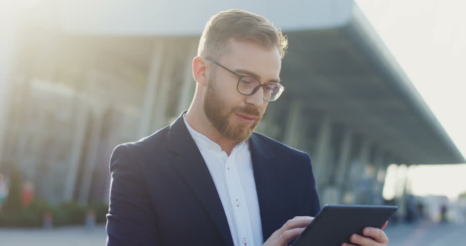 Close up of the attractive young businessman taping and scrolling on the tablet computer, then turning his head and smiling to the camera. Portrait. Outdoor.