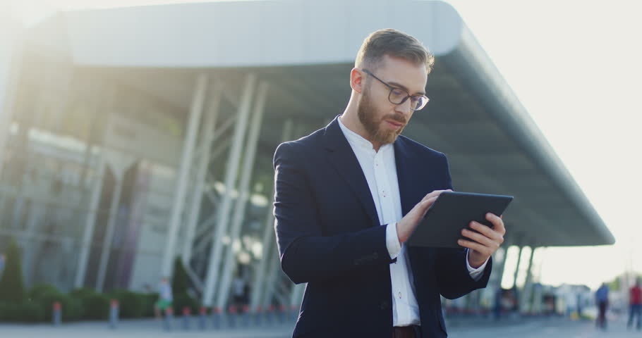 Portrait shot of the Caucasian young office worker in the glasses using his tablet computer - scrolling, taping and texting while standing near the big modern building, airport or train station. | Shutterstock HD Video #1014757736