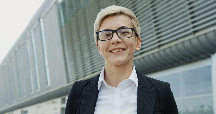 Portrait of the succesful middle aged businesswoman with short blond hair turning her head to the camera and smiling on the big urban building background. Close up