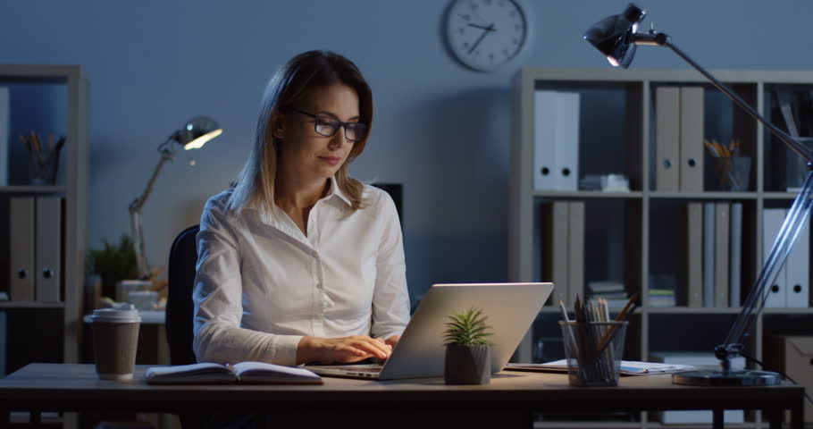 Caucasian female office worker still being late at night in the office at the laptop computer, trying to rest while massaging her neck.
