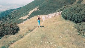 Aerial View Running woman on the top of mountain. Runner is jogging in summer sunny day around trees and mountains background under sun light with blue sky, Drone 4K Video
