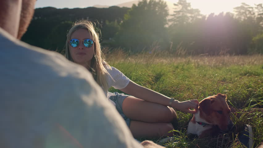 young woman talking to friends at picnic while petting her dog, sun flare in background