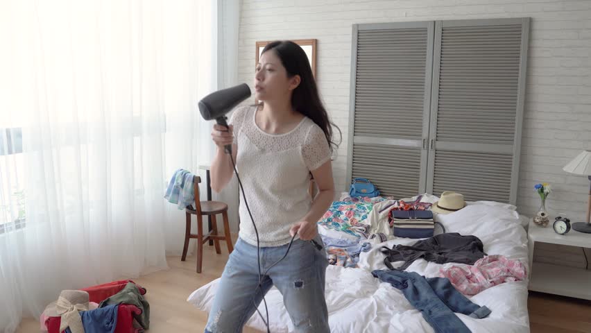 Slow motion of the young modern Asian woman singing in her bedroom using the hairdryer. She sinks herself into the pop music. She is imitating the superstars. Royalty-Free Stock Footage #1014776258