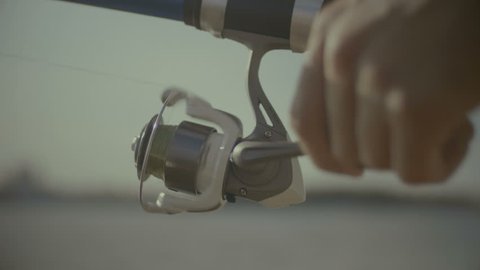 Closeup of fisherman hands holding fishing rod and twisting coil while fishing. Male hands with fishing rod twisting spinning reel while catching a fish on sunny summer day. Slow motion.