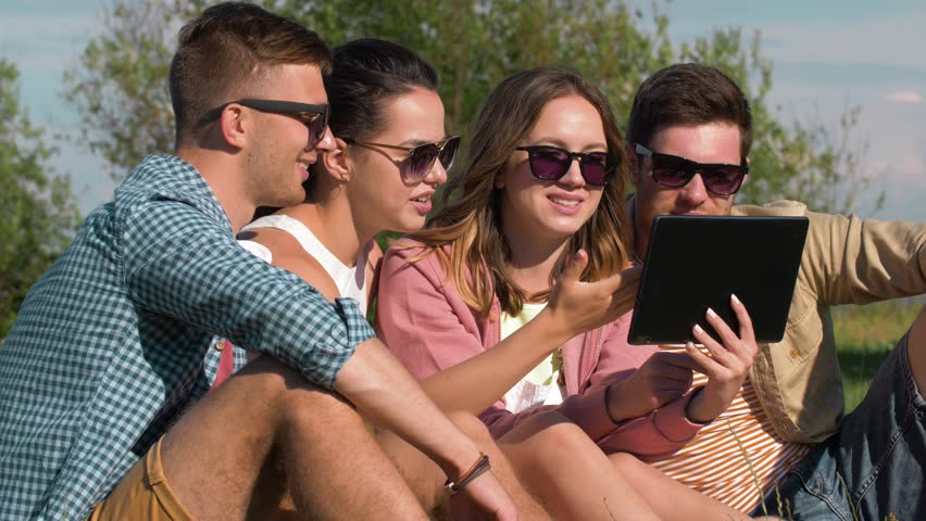 friendship, technology and leisure concept - group of smiling friends with tablet pc computer sitting on grass at summer park