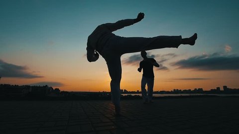 Silhouettes of two sports men who train skills of martial art of capoeira against the background of the amazing sky
