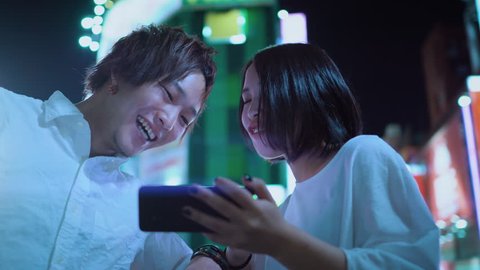 Young East Asian Boy and Girl Couple Talking, Laughing, Using Mobile Phone and Sharing Screen. In the Background Blurred Advertising Billboards and City Lights at Night. 
