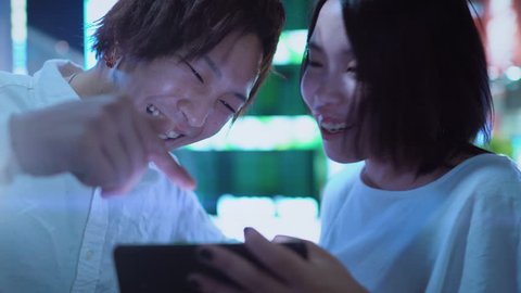 Young Japanese Boy and Girl Couple Talking, Laughing, Using Mobile Phone and Sharing Screen. In the Background Blurred Advertising Billboards and City Lights at Night. 