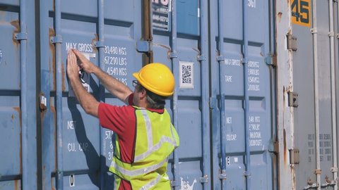 Shipping worker sticking a biohazard sign on a shipping container in a shipping yard filled with large shipping containers.