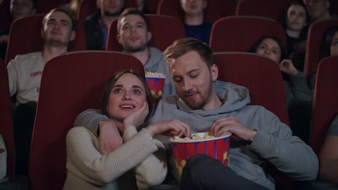 Love couple watching movie in cinema theatre. Couple in love embracing in cinema. People enjoying film and eating popcorn at cinema in slow motion. Movie entertainment