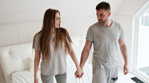 Handheld shot of happy young couple in sleepwear holding hands and standing on bed, then falling down on mattress and laughing