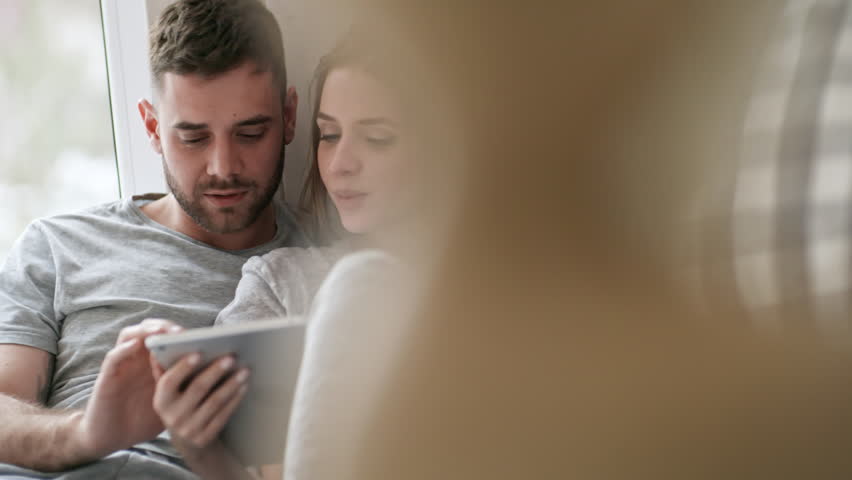Handheld shot of cheerful young woman and bearded man in sleepwear sitting on bed and chatting while using tablet in morning