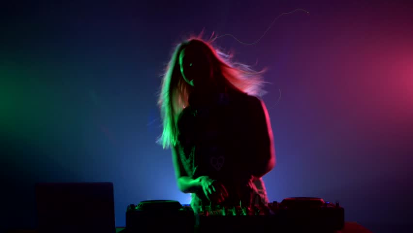 Young, smiling, sexy, attractive dj girl in sexy clothes, flipping hair, hands up and enjoy it, using headphones, smoke, green, blue and pink backlight, silhouette