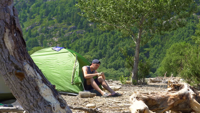 A tourist, sitting in a tent located on the shore of a mountain lake, carves a wood article.