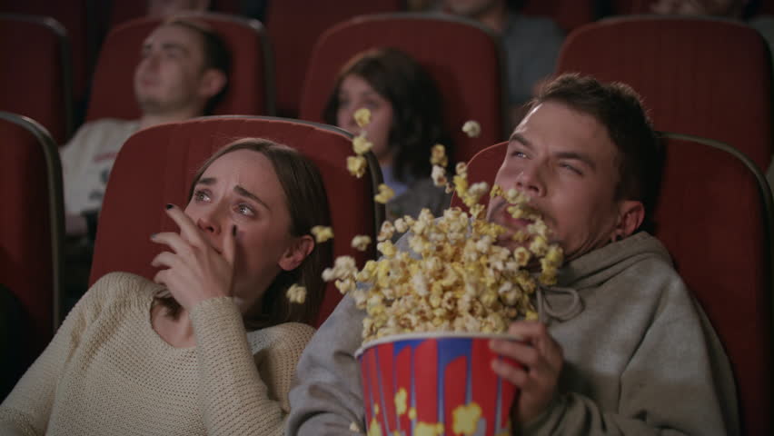 Young couple watching horror film in movie theatre. Guy sprinkle popcorn on girl in movie theater. Scared guy jumping in seat and spilling popcorn in slow motion. Shocked people watching horror movie | Shutterstock HD Video #1014864670