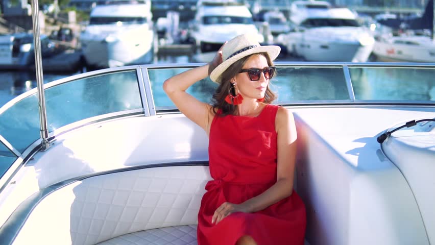 Young girl in red dress, hat and sunglasses sitting on a chair in a white yacht on the background of water and boats