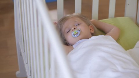 A little girl is sleeping in a baby crib with a pacifier in her mouth