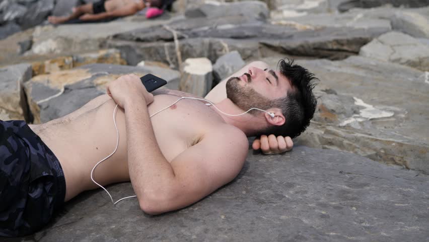 Solitary fit young man laying on a stone at the beach while listening to music through earphones
