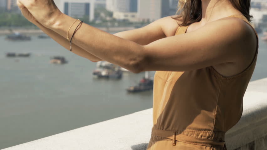 Young woman taking photos of cityscape with cellphone standing on terrace in city
