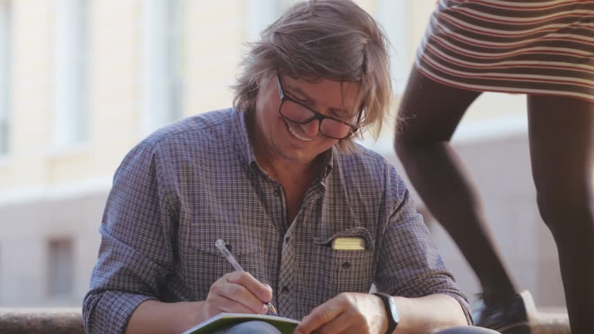 Middle-aged european man in eyeglasses casual dressed writing down notes in his copybook, smiling, drinking coffee sitting on stairs outdoors.