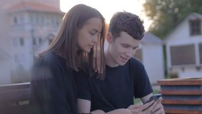Couple sharing media in a smart phone sitting in a bench in a park with buildings in the background