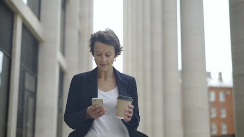 Medium dolly shot of elegant middle aged businesswoman with short hair walking down street with cell phone and takeaway coffee cup in her hands and looking around thoughtfully
