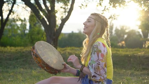 a blond hippie woman playing a large leather tambourine and singing. Attractive girl with braids resting in the park at sunset. sits on a veil on green grass.