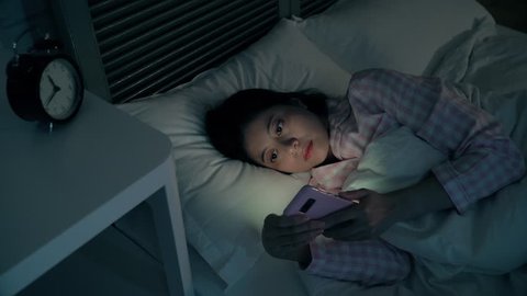 Modern young lady lying on the bed and using texting on the smartphone in the dark night. She is so lonely and tries to get on the social website to find someone to chat with.