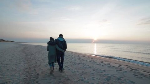 Young beautiful enamored couple walking slowly side by side along coastline on the beach with wet sand by the sea, watching sunset and talking
