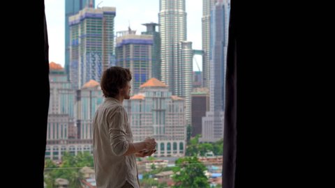 Young man comes to a balcony of his apartment with a view on a city centre full of skyscrappers and drinks his morning coffee