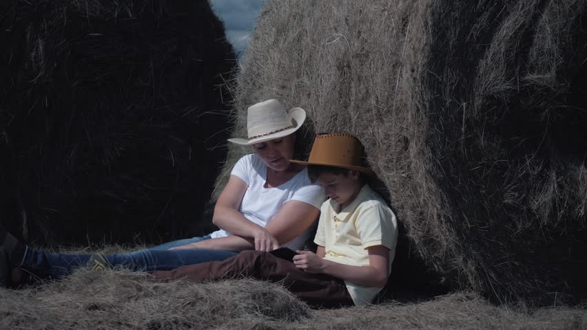 Happy family mother and son with a tablet sitting in the field, leaning on a hay-roll. The concept of a happy family