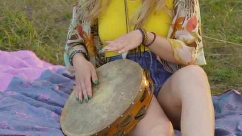 young blond hippie woman sitting on grass in park at sunset. Warm summer day and soft backlight. happy woman playing the tambourine and singing.