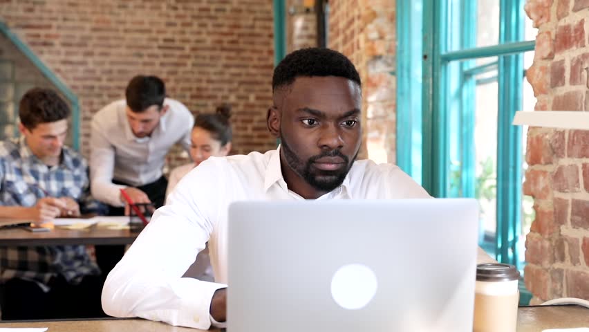 Male Afroamerican Working at the Office. Sitting at the Table with Modern Computer. Looking at the Screen, Camera. Smiling Sincerly. Young Man Working on Start up. Business People in the Background.