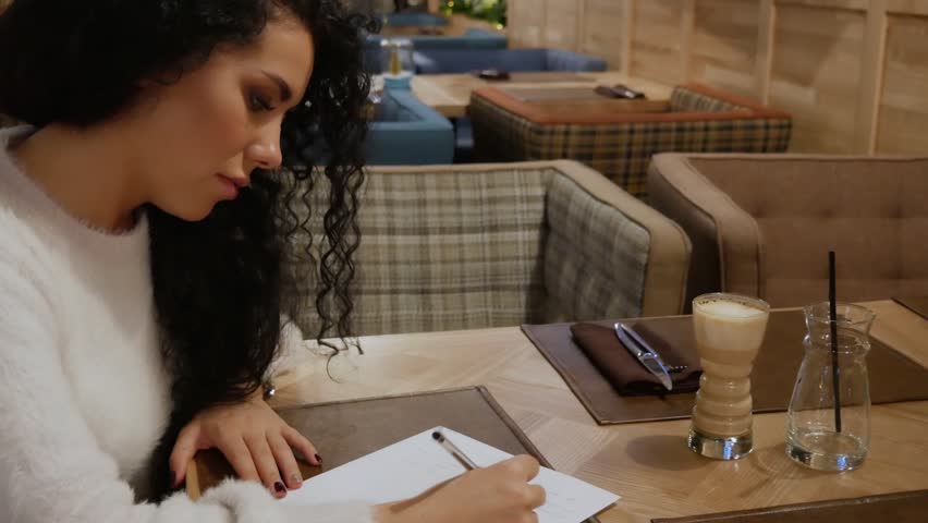 Cute girl with curly hair is writing a poem in a restaurant. Brunette writer in a white sweater creates novel at winter cafe