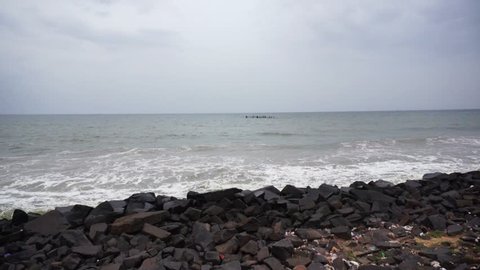 Left to right pan of a rocky beach with black rocks and a choppy sea and cloudy sky shot at evening in pondicherry. This famed vacation spot is a couple of hours drive away from chennai