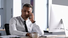 Displeased serious man in white shirt talking on phone while working in office