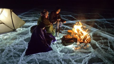 Three travelers by fire right on ice at night. Campground on ice. Tent stands next to fire. Lake Baikal. Nearby there is car. People are warming around campfire and are dressed in sleeping bags. This 库存视频