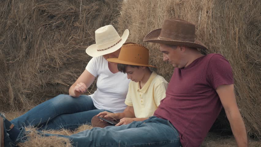 Happy family mother, father, and son with a tablet sitting in the field, leaning on a hay-roll. The concept of a happy family