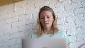 Attractive young woman with long blond hair wearing a green blue t shirt sitting against a white brick wall with her laptop and video chatting. Slider slow motion medium low angle shot