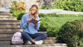 Cheerful young blonde lady sitting in park and talking on smartphone while reading book