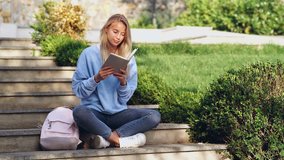 Cocentrated smiling blonde lady sitting in park and reading book while studying