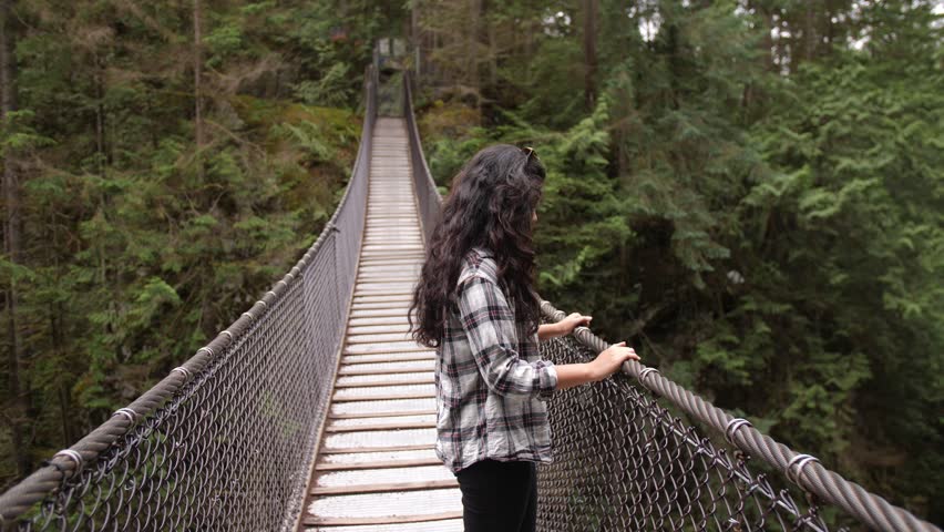 Dark Haired Latina Woman Leaning Over Side of Suspension Bridge Appearing Astonished and Happy Pointing at Nearby Waterfall Wearing Checkered Shirt in Thick Canadian Forest | Shutterstock HD Video #1015059376