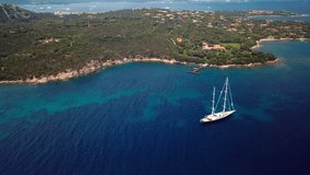 Video from above, aerial view of a luxury sailboat at anchor on a transparent and turquoise sea in front of a green coast of Sardinia, Italy.