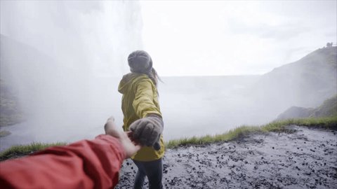 Young couple on a hike in Iceland. Couple holding hands: woman leading boyfriends walking towards waterfall POV travel and adventure concept. Follow me to. 